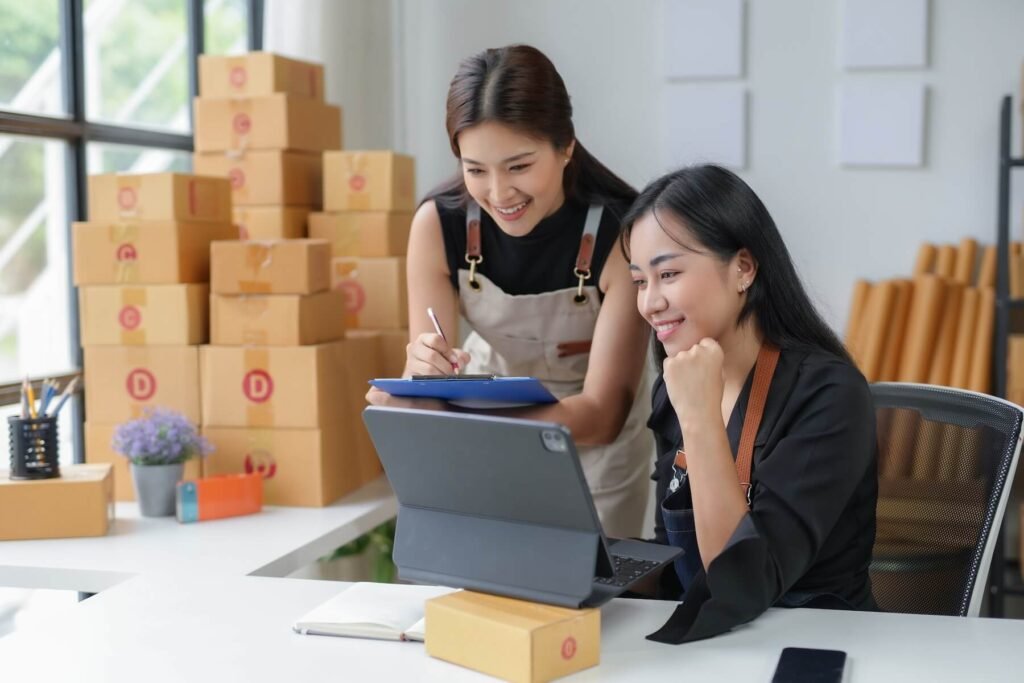 Two small business owners checking online orders on a tablet amid shipping boxes